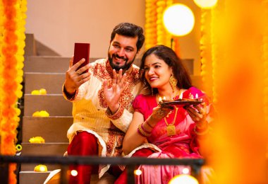 Diwali 'ye video çağrısı yapan çift, Saree' de Hintli Genç Evli Çifti ve Şenlik Gecesinde Smartphone aracılığıyla Akrabalarla Gülümseyen Diya Thali 'yi tutan süslü merdivenlerde oturan Kurta