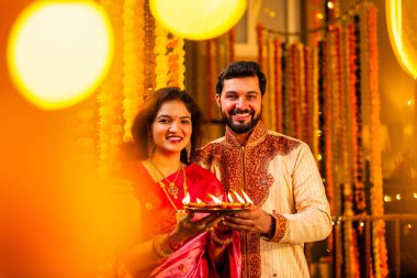 Geleneksel Saree 'deki Hintli evli çift Diya Thali ve Marigold Garland' a Karşı Ayakta Duran, Terracotta Petrol Lambası Tabağı Tutan ve Festivalde Kamerada Gülümseyen