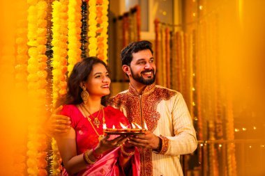 Geleneksel Saree 'deki Hintli evli çift Diya Thali ve Marigold Garland' a Karşı Ayakta Duran, Terracotta Petrol Lambası Tabağı Tutan ve Festivalde Kamerada Gülümseyen