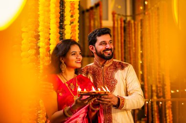 Geleneksel Saree 'deki Hintli evli çift Diya Thali ve Marigold Garland' a Karşı Ayakta Duran, Terracotta Petrol Lambası Tabağı Tutan ve Festivalde Kamerada Gülümseyen