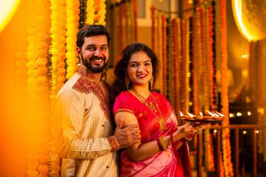 Geleneksel Saree 'deki Hintli evli çift Diya Thali ve Marigold Garland' a Karşı Ayakta Duran, Terracotta Petrol Lambası Tabağı Tutan ve Festivalde Kamerada Gülümseyen