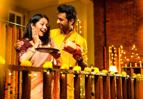 Karı koca Diwali kutlaması sırasında hediye veriyor, Hintli çift geleneksel kıyafet hediye ediyor ve evdeki ahşap korkuluklara gaz lambası yerleştiriyor, parıltılı diya ve dekorasyonlarla şenlikli bir akşamın tadını çıkarıyor.