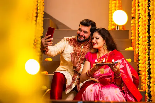 Diwali 'ye video çağrısı yapan çift, Saree' de Hintli Genç Evli Çifti ve Şenlik Gecesinde Smartphone aracılığıyla Akrabalarla Gülümseyen Diya Thali 'yi tutan süslü merdivenlerde oturan Kurta