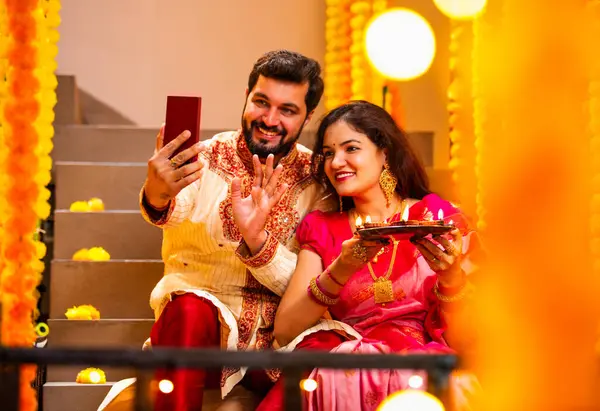 Diwali 'ye video çağrısı yapan çift, Saree' de Hintli Genç Evli Çifti ve Şenlik Gecesinde Smartphone aracılığıyla Akrabalarla Gülümseyen Diya Thali 'yi tutan süslü merdivenlerde oturan Kurta