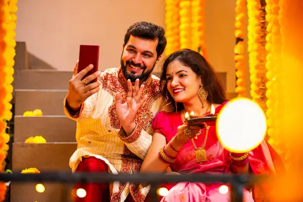 Diwali 'ye video çağrısı yapan çift, Saree' de Hintli Genç Evli Çifti ve Şenlik Gecesinde Smartphone aracılığıyla Akrabalarla Gülümseyen Diya Thali 'yi tutan süslü merdivenlerde oturan Kurta