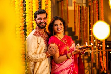 Geleneksel Saree 'deki Hintli evli çift Diya Thali ve Marigold Garland' a Karşı Ayakta Duran, Terracotta Petrol Lambası Tabağı Tutan ve Festivalde Kamerada Gülümseyen