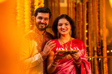 Geleneksel Saree 'deki Hintli evli çift Diya Thali ve Marigold Garland' a Karşı Ayakta Duran, Terracotta Petrol Lambası Tabağı Tutan ve Festivalde Kamerada Gülümseyen