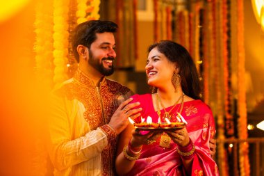 Geleneksel Saree 'deki Hintli evli çift Diya Thali ve Marigold Garland' a Karşı Ayakta Duran, Terracotta Petrol Lambası Tabağı Tutan ve Festivalde Kamerada Gülümseyen