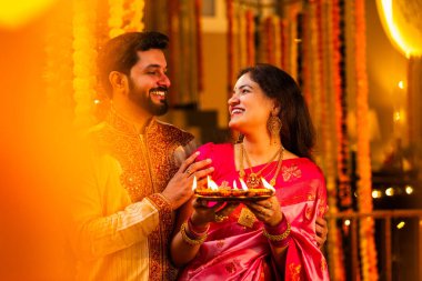 Geleneksel Saree 'deki Hintli evli çift Diya Thali ve Marigold Garland' a Karşı Ayakta Duran, Terracotta Petrol Lambası Tabağı Tutan ve Festivalde Kamerada Gülümseyen