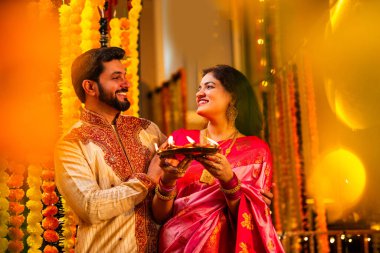 Geleneksel Saree 'deki Hintli evli çift Diya Thali ve Marigold Garland' a Karşı Ayakta Duran, Terracotta Petrol Lambası Tabağı Tutan ve Festivalde Kamerada Gülümseyen