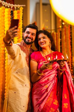 Geleneksel Kurta ve Saree Standing Against Marigold Çelenkleri 'nde gülümseyen Hintli genç çift Diya Thali' yi tutarken video çağrısı.