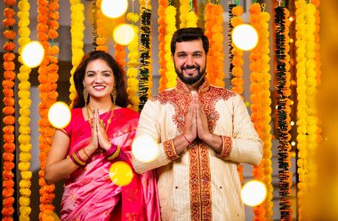 Katlanmış Elleri ve Eğilimi olan çift, Geleneksel Giyinen Hintli Çift Namaste Jestleriyle Kameraya Bakıyor, Diwali Sırasındaki Marigold Garland 'ın önünde Gülümsüyor