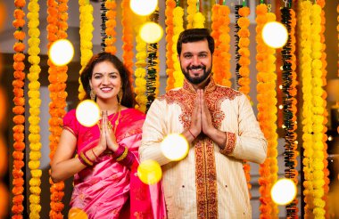 Katlanmış Elleri ve Eğilimi olan çift, Geleneksel Giyinen Hintli Çift Namaste Jestleriyle Kameraya Bakıyor, Diwali Sırasındaki Marigold Garland 'ın önünde Gülümsüyor