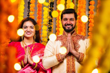 Katlanmış Elleri ve Eğilimi olan çift, Geleneksel Giyinen Hintli Çift Namaste Jestleriyle Kameraya Bakıyor, Diwali Sırasındaki Marigold Garland 'ın önünde Gülümsüyor