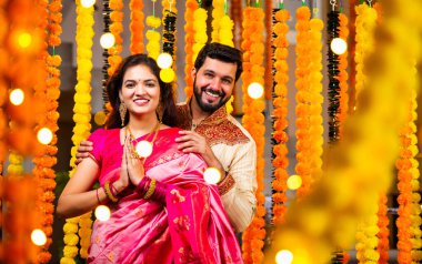 Katlanmış Elleri ve Eğilimi olan çift, Geleneksel Giyinen Hintli Çift Namaste Jestleriyle Kameraya Bakıyor, Diwali Sırasındaki Marigold Garland 'ın önünde Gülümsüyor