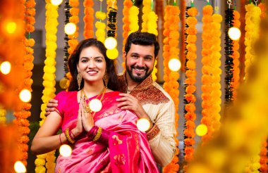 Katlanmış Elleri ve Eğilimi olan çift, Geleneksel Giyinen Hintli Çift Namaste Jestleriyle Kameraya Bakıyor, Diwali Sırasındaki Marigold Garland 'ın önünde Gülümsüyor