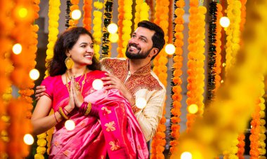 Katlanmış Elleri ve Eğilimi olan çift, Geleneksel Giyinen Hintli Çift Namaste Jestleriyle Kameraya Bakıyor, Diwali Sırasındaki Marigold Garland 'ın önünde Gülümsüyor