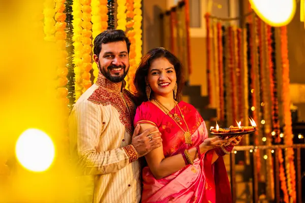 Geleneksel Saree 'deki Hintli evli çift Diya Thali ve Marigold Garland' a Karşı Ayakta Duran, Terracotta Petrol Lambası Tabağı Tutan ve Festivalde Kamerada Gülümseyen