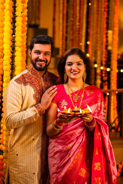 Geleneksel Saree 'deki Hintli evli çift Diya Thali ve Marigold Garland' a Karşı Ayakta Duran, Terracotta Petrol Lambası Tabağı Tutan ve Festivalde Kamerada Gülümseyen