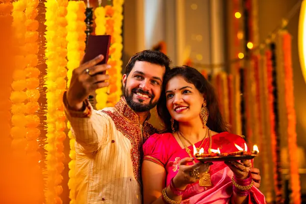 Geleneksel Kurta ve Saree Standing Against Marigold Çelenkleri 'nde gülümseyen Hintli genç çift Diya Thali' yi tutarken video çağrısı.
