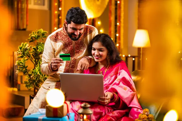Online Diwali Alışveriş yapmak Laptop ve Kredi Kartı kullanmak. Geleneksel Etnik Giyimli Şenlik Kutlamaları sırasında Tatlılarla Süslü Ev 'de oturan Hintli genç çift
