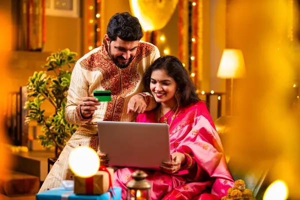 Online Diwali Alışveriş yapmak Laptop ve Kredi Kartı kullanmak. Geleneksel Etnik Giyimli Şenlik Kutlamaları sırasında Tatlılarla Süslü Ev 'de oturan Hintli genç çift
