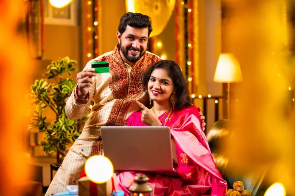 Online Diwali Alışveriş yapmak Laptop ve Kredi Kartı kullanmak. Geleneksel Etnik Giyimli Şenlik Kutlamaları sırasında Tatlılarla Süslü Ev 'de oturan Hintli genç çift