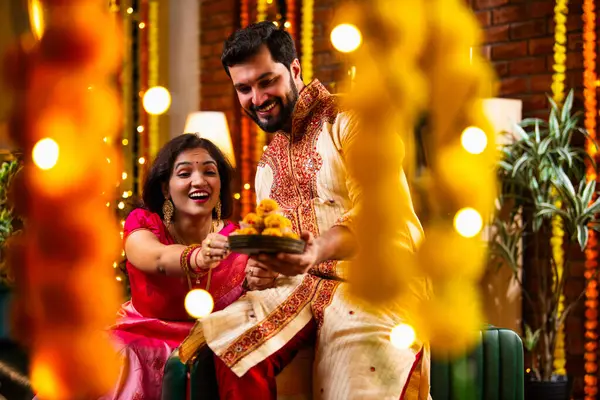 Tatlılar, Hediye Kutuları, Diwali Dekorasyonları, Diwali Festivali gecesi kanepede oturup Neşeyle Gülen Çiftler Birbirlerinden Tatlı Laddu Kapıyor