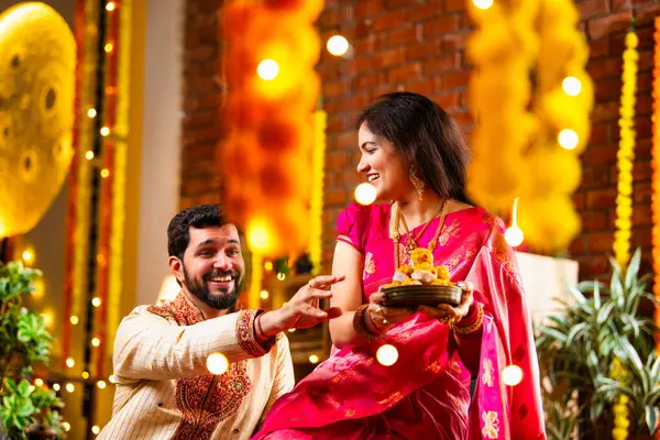 Tatlılar, Hediye Kutuları, Diwali Dekorasyonları, Diwali Festivali gecesi kanepede oturup Neşeyle Gülen Çiftler Birbirlerinden Tatlı Laddu Kapıyor