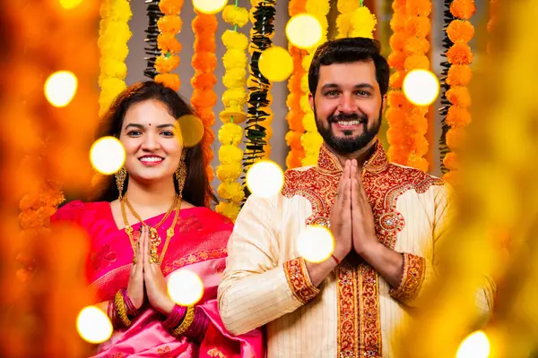 Katlanmış Elleri ve Eğilimi olan çift, Geleneksel Giyinen Hintli Çift Namaste Jestleriyle Kameraya Bakıyor, Diwali Sırasındaki Marigold Garland 'ın önünde Gülümsüyor