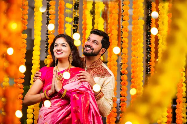 Katlanmış Elleri ve Eğilimi olan çift, Geleneksel Giyinen Hintli Çift Namaste Jestleriyle Kameraya Bakıyor, Diwali Sırasındaki Marigold Garland 'ın önünde Gülümsüyor