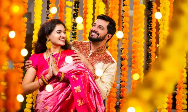 Katlanmış Elleri ve Eğilimi olan çift, Geleneksel Giyinen Hintli Çift Namaste Jestleriyle Kameraya Bakıyor, Diwali Sırasındaki Marigold Garland 'ın önünde Gülümsüyor