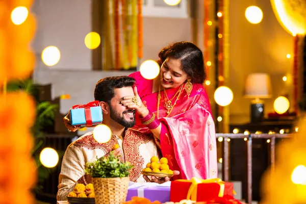 Geleneksel Şenlik Giysileriyle Evde Otururken, Evde Işıklar ve Marigold çelenkleriyle süslenmiş Diwali Gecesini Gülümseyen ve Kutlayan Hintli Eş Kocaya Sürpriz Hediye Veriyor