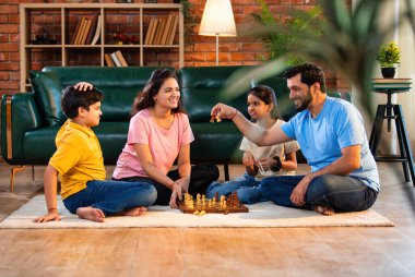 Hintli aile satranç oynuyor. Halı döşemesinde şık bir oturma odasında oturuyorlar. Çocuklar ve ebeveynlerle evde vakit geçirip hafta sonu aktivitelerinin keyfini çıkarıyorlar.