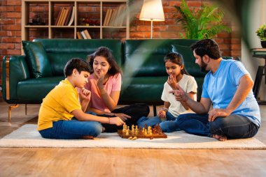 Hintli aile satranç oynuyor. Halı döşemesinde şık bir oturma odasında oturuyorlar. Çocuklar ve ebeveynlerle evde vakit geçirip hafta sonu aktivitelerinin keyfini çıkarıyorlar.