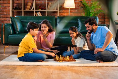 Hintli aile satranç oynuyor. Halı döşemesinde şık bir oturma odasında oturuyorlar. Çocuklar ve ebeveynlerle evde vakit geçirip hafta sonu aktivitelerinin keyfini çıkarıyorlar.