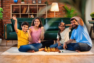 Hintli aile satranç oynuyor. Halı döşemesinde şık bir oturma odasında oturuyorlar. Çocuklar ve ebeveynlerle evde vakit geçirip hafta sonu aktivitelerinin keyfini çıkarıyorlar.