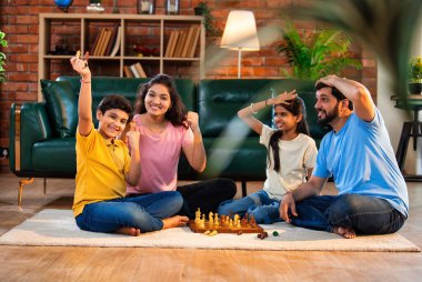 Hintli aile satranç oynuyor. Halı döşemesinde şık bir oturma odasında oturuyorlar. Çocuklar ve ebeveynlerle evde vakit geçirip hafta sonu aktivitelerinin keyfini çıkarıyorlar.