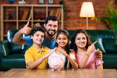 Çocuklar domuz kumbarasında para biriktirirken Hintli aileler arka planda gülümsüyor, mutlu oğul ve kız para kutusuna bozuk para koyuyorlar, evde rahat oturma odasında mali alışkanlıklar öğreniyorlar.