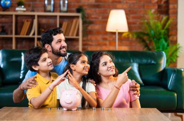 Çocuklar domuz kumbarasında para biriktirirken Hintli aileler arka planda gülümsüyor, mutlu oğul ve kız para kutusuna bozuk para koyuyorlar, evde rahat oturma odasında mali alışkanlıklar öğreniyorlar.