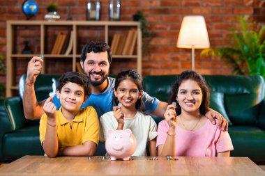 Çocuklar domuz kumbarasında para biriktirirken Hintli aileler arka planda gülümsüyor, mutlu oğul ve kız para kutusuna bozuk para koyuyorlar, evde rahat oturma odasında mali alışkanlıklar öğreniyorlar.