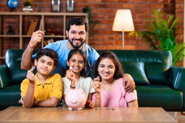 Çocuklar domuz kumbarasında para biriktirirken Hintli aileler arka planda gülümsüyor, mutlu oğul ve kız para kutusuna bozuk para koyuyorlar, evde rahat oturma odasında mali alışkanlıklar öğreniyorlar.