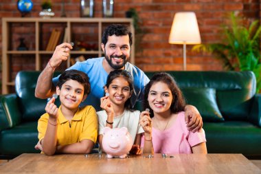 Çocuklar domuz kumbarasında para biriktirirken Hintli aileler arka planda gülümsüyor, mutlu oğul ve kız para kutusuna bozuk para koyuyorlar, evde rahat oturma odasında mali alışkanlıklar öğreniyorlar.