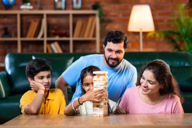 Hintli ebeveynler küçük çocuklarla vakit geçirmenin keyfini çıkarırken ve rahat modern oturma odasında ahşap blokları dikkatlice dengelerken aile evde ahşap blok oyunu oynuyor.