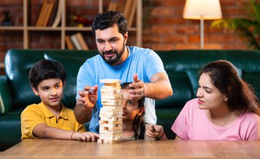 Hintli ebeveynler küçük çocuklarla vakit geçirmenin keyfini çıkarırken ve rahat modern oturma odasında ahşap blokları dikkatlice dengelerken aile evde ahşap blok oyunu oynuyor.