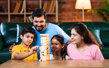 Hintli ebeveynler küçük çocuklarla vakit geçirmenin keyfini çıkarırken ve rahat modern oturma odasında ahşap blokları dikkatlice dengelerken aile evde ahşap blok oyunu oynuyor.