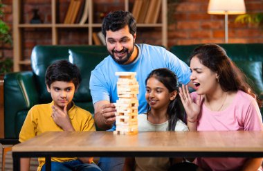 Hintli ebeveynler küçük çocuklarla vakit geçirmenin keyfini çıkarırken ve rahat modern oturma odasında ahşap blokları dikkatlice dengelerken aile evde ahşap blok oyunu oynuyor.