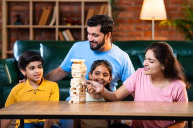 Hintli ebeveynler küçük çocuklarla vakit geçirmenin keyfini çıkarırken ve rahat modern oturma odasında ahşap blokları dikkatlice dengelerken aile evde ahşap blok oyunu oynuyor.