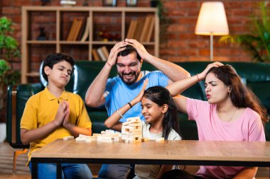 Hintli ebeveynler küçük çocuklarla vakit geçirmenin keyfini çıkarırken ve rahat modern oturma odasında ahşap blokları dikkatlice dengelerken aile evde ahşap blok oyunu oynuyor.