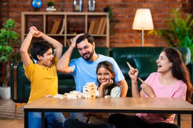 Hintli ebeveynler küçük çocuklarla vakit geçirmenin keyfini çıkarırken ve rahat modern oturma odasında ahşap blokları dikkatlice dengelerken aile evde ahşap blok oyunu oynuyor.